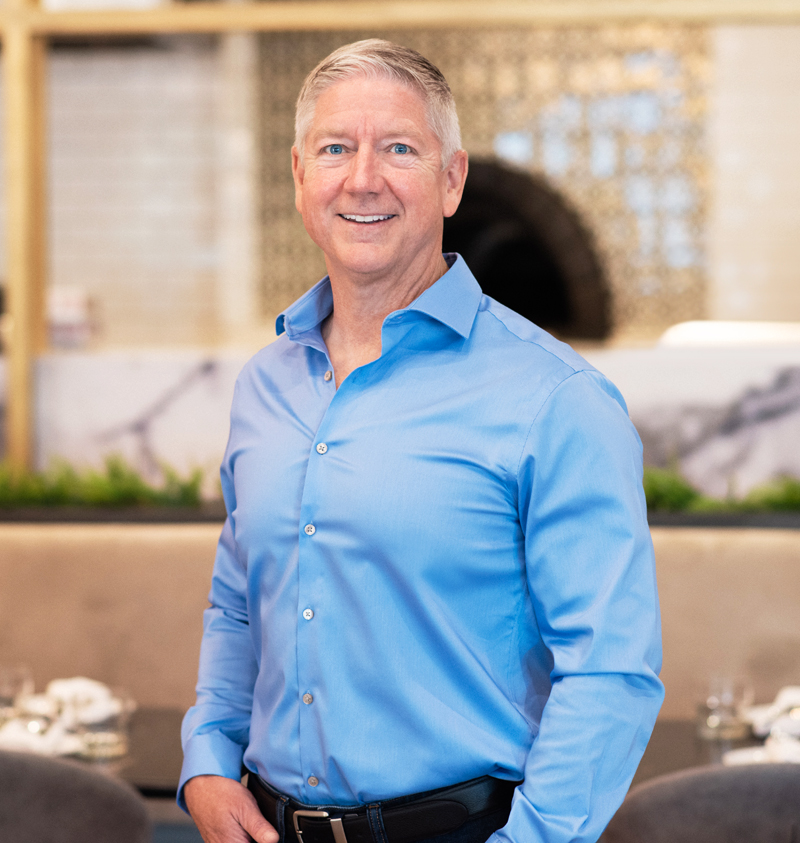 Portrait of Paul Munsterman, smiling and wearing a light blue dress shirt, standing in a modern restaurant interior with a neutral and elegant background.